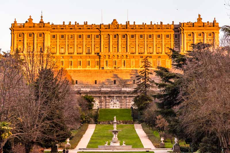 Winter golden hour photograph of Madrid Royal Palace with English garden with fountain. Sunset colours on Baroque façade.